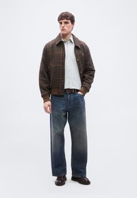 Νεαρός άνδρας που φοράει καφέ καρό σακάκι, ανοιχτό γκρι πουκάμισο με κουμπιά, μπλε τζιν φαρδύ, και σκούρα καφέ παπούτσια, στέκεται μπροστά σε λευκό φόντο.