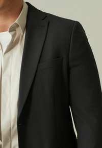 Sort blazer med skræddersyet pasform, notch lapels, enkelt knaplukning og brystlomme. Båret over en hvid skjorte. Glat tekstur.