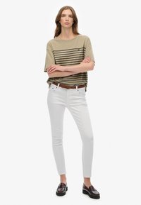 Olivenstribet kortærmet skjorte, hvide skinny jeans, brun flettet bælte og sorte loafers. Modellen står med armene foldet, neutral baggrund.