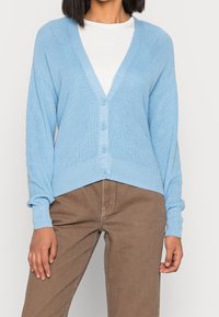 Ljusblå stickad kofta med V-hals, fyra knappar och ribbad textur. Har drop shoulders och små öppna mönster nära axlarna.