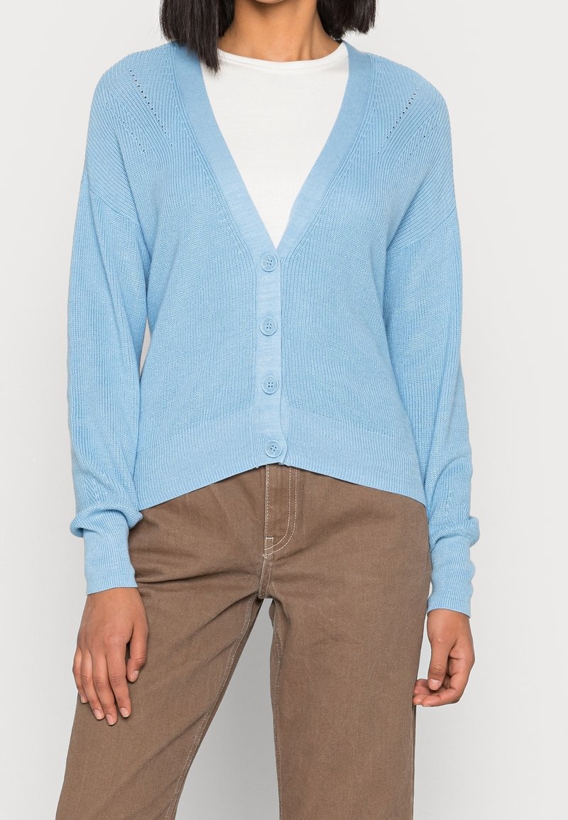 Ljusblå stickad kofta med V-hals, fyra knappar och ribbad textur. Har drop shoulders och små öppna mönster nära axlarna.