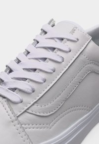 Baskets en cuir blanc avec lacets blancs, détail de couture latéral, et une semelle profilée basse, montrées en gros plan.