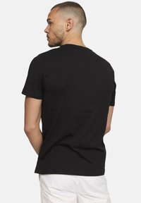 Homme aux cheveux courts et à la barbe, portant un t-shirt noir et un pantalon blanc, de dos et regardant vers la gauche sur fond uni.