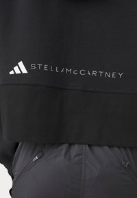 Černá zkrácená mikina s ribovanou texturou. Na přední straně se nachází bílé logo "adidas" a text "STELLA McCARTNEY".