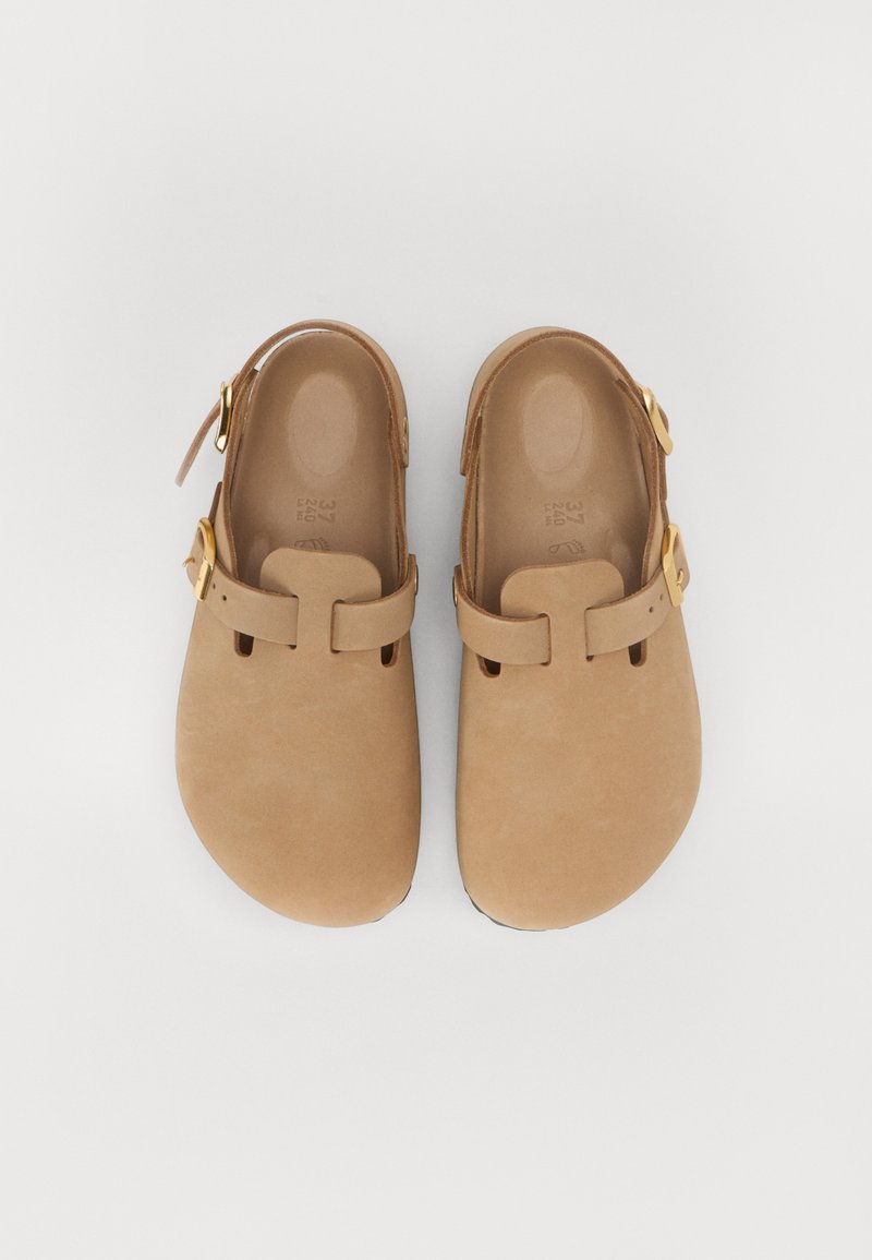 Zoccoli marroni con una finitura liscia e opaca, dotati di una fascia sicura sul collo del piede e un accento in fibbia dorata. Design con punta arrotondata.