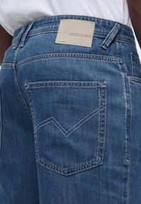 Κοντινό πλάνο πίσω τσέπης μπλε τζιν με ραμμένο σχέδιο και μπεζ ετικέτα με το όνομα της μάρκας πάνω από τη μέση.