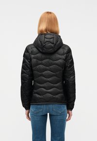 Άτομο με μαλλιά έως τους ώμους, φορώντας μαύρο καπιτονέ μπουφάν με κουκούλα και μπλε τζιν, στέκεται με την πλάτη γυρισμένη σε απλό φόντο.