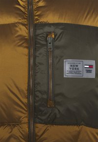 Steppjacke in Braun und Dunkelgrün. Mit durchgehendem Reißverschluss, kontrastierenden Stoffen und einem Logo-Patch mit Schriftzug. Glatte, glänzende Oberfläche.