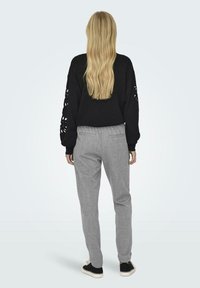 Schwarzes, kurzes Sweatshirt mit Ausschnitten an den Ärmeln, kombiniert mit hellgrauen gemusterte Hosen und schwarzen Turnschuhen.