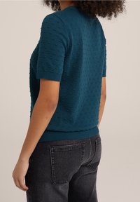 Pull manches courtes turquoise avec motif à boucles texturé, col et poignets côtelés, associé à un jean gris foncé.