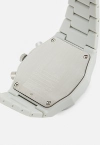 Montre avec bracelet en silicone gris et dos en acier inoxydable brossé affichant des marquages et spécifications. Présente un design élégant et angulaire.