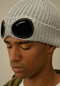 Beanie grigio lavorato a maglia con grandi occhiali neri dal finish lucido. È visibile un motivo testurizzato e un bordo arrotolato.