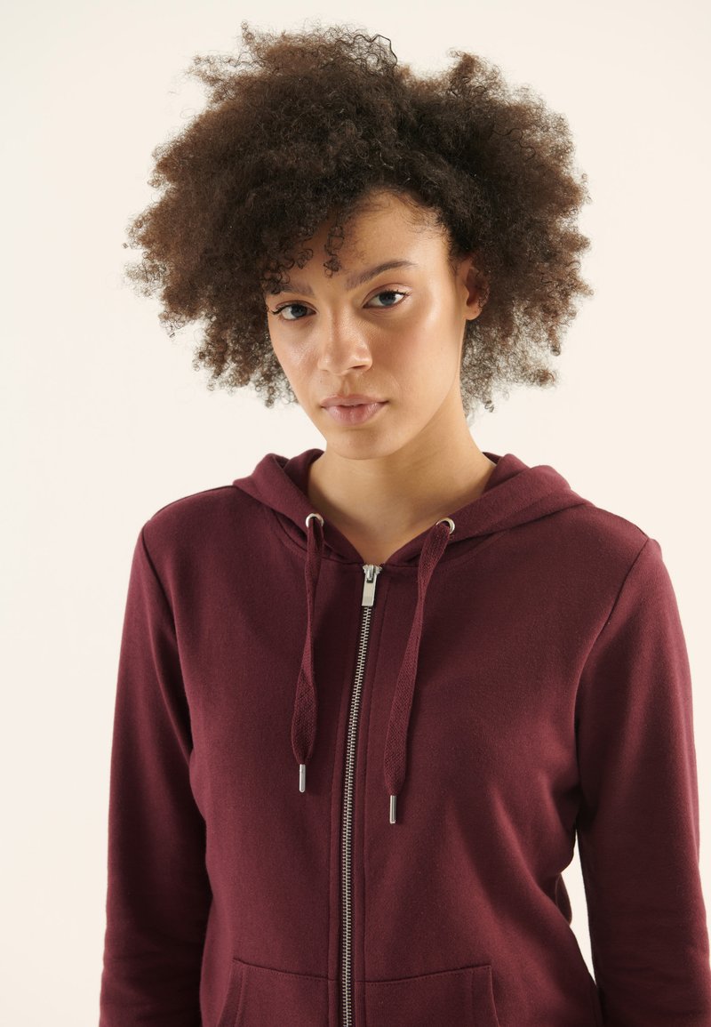 Mlada ženska s kodrastimi lasmi, oblečena v vinasto rdečo kapuco z zadrgo in vrvicami, obrnjena proti kameri na enostavni svetli podlagi.