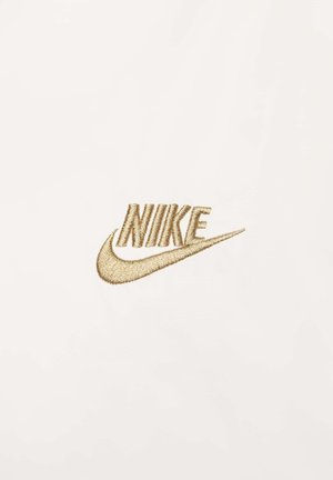 Logo Nike en broderie dorée sur un fond en tissu blanc texturé, avec des lettres stylisées et un design de swoosh distinctif.