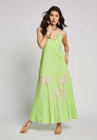 Grünes Maxikleid mit tiefem V-Ausschnitt, verziert mit rosa bestickten Palmen am Saum und einem ausgefransten Rand. Transparente Sandalen runden das Outfit ab.
