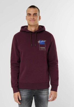Maroon hoodie with a front pocket, drawstring hood, ribbed cuffs and hem, featuring a graphic patch with "Explore" text. Worn with gray jeans.