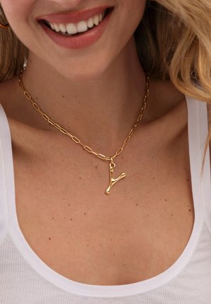 Mujer sonriente con un collar de cadena dorada con un colgante dorado en forma de Y y una camiseta sin mangas blanca.