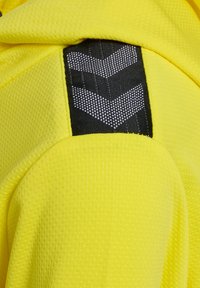 Gul atletisk hoodie i tekstureret stof med et fremtrædende sort patch. Sort chevron-mønster med refleksdetaljer.