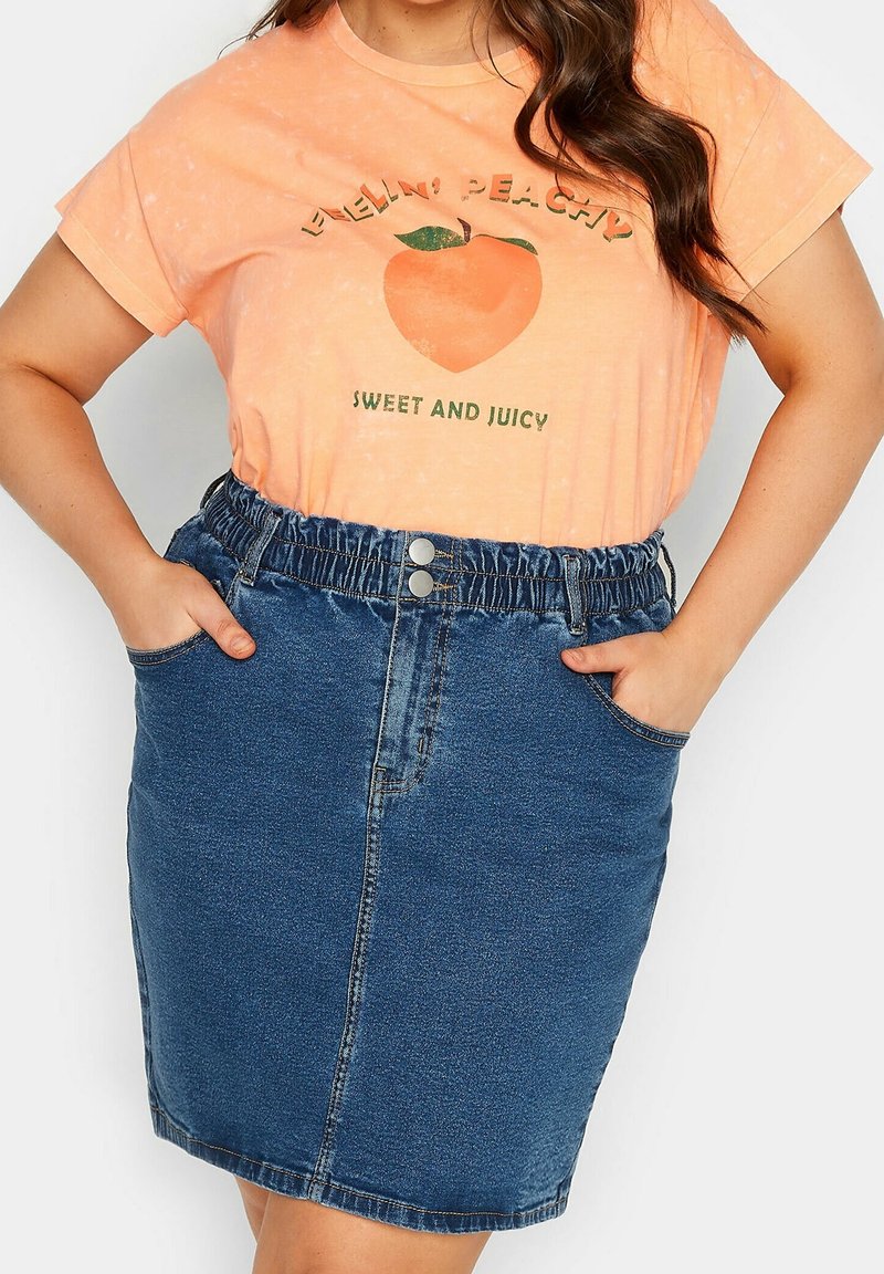 Tričko meruňkové barvy s grafickým potiskem v kombinaci s tmavými džínovými sukněmi se strečovým pasem a dvěma bočními kapsami.