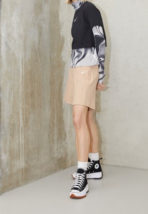 Persoon die een zwart-wit gelaagde top, beige Nike-shorts, witte sokken en zwarte hoge sneakers draagt, staand tegen een betonnen muur.