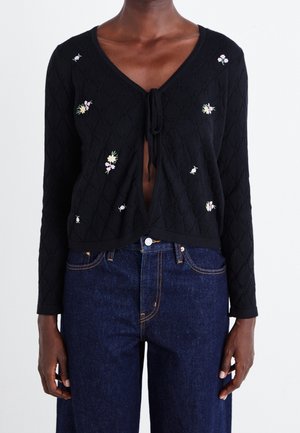 Cardigan noir à manches longues en maille avec petite broderie florale, noué sur la poitrine, porté avec un jean taille haute bleu foncé.
