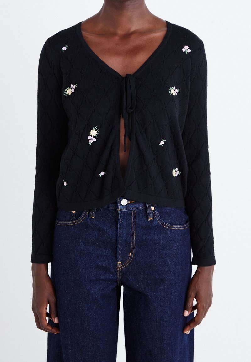 Cardigan noir à manches longues en maille avec petite broderie florale, noué sur la poitrine, porté avec un jean taille haute bleu foncé.