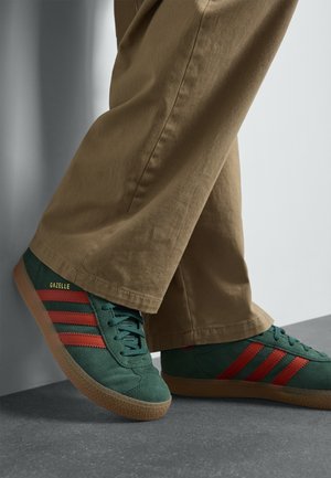 Grønne ruskindssneakers med røde striber og gul "GAZELLE"-branding. Skoene har en sål i gummigummi og er kombineret med beige bukser.