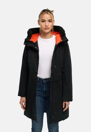 Vrouw die een zwarte winterjas met capuchon en oranje voering draagt, wit overhemd en blauwe jeans, staand tegen een effen achtergrond.