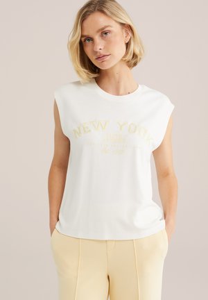 Blonde Frau trägt ein ärmelloses weißes Oberteil mit dem Schriftzug "New York" und hellbeige Hosen, steht vor einem schlichten neutralen Hintergrund.