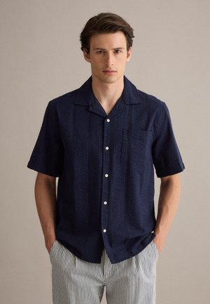 Joven con una camisa de manga corta azul marino texturizada con botones blancos y pantalones claros a rayas, de pie con las manos en los bolsillos.