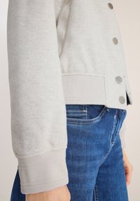 Hellgraue Fleecejacke mit einer strukturierten Oberfläche, ausgestattet mit Druckknöpfen und gerippten Bündchen, kombiniert mit blauen Jeans.