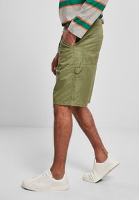 Person som bär olivgröna cargo-shorts, vita sneakers och en långärmad skjorta med gröna och orange ränder, stående mot en enkel bakgrund.