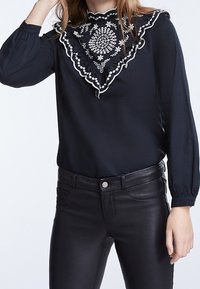 Sort bluse med en flæsekant i halsen, prydet med hvid blomsterbroderi, kombineret med tætsiddende sorte læderbukser. Lange ærmer med samlet manchet.