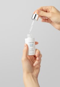 Witte serumfles met het etiket "TIRTIR PHA 15% SERUM", vastgehouden door een hand. De druppelaar haalt een heldere vloeistof eruit, met een glanzend zilveren dop.