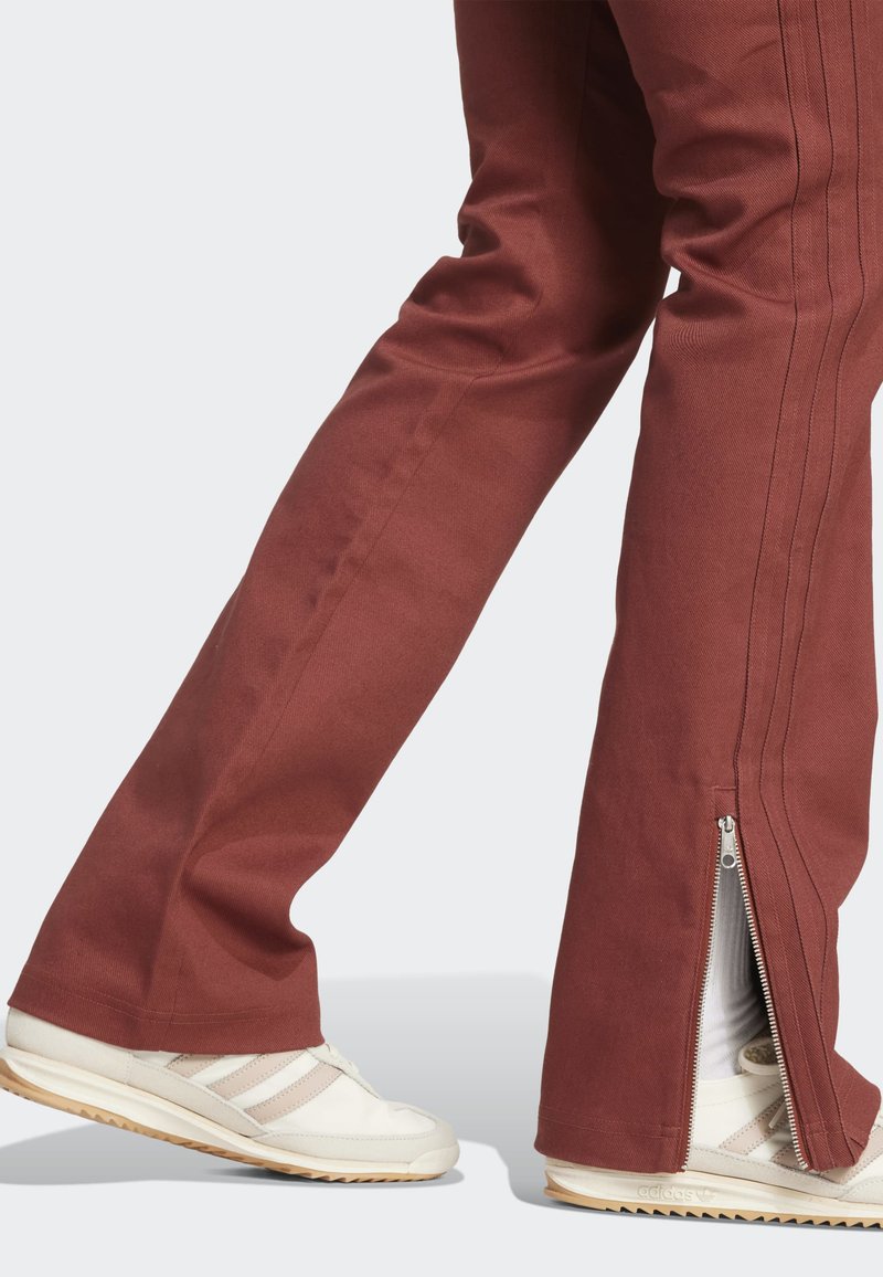 Vue en longueur des jambes d'une personne portant un pantalon couleur rouille avec des fermetures éclair latérales et des baskets Adidas blanches avec des bandes beige.