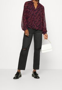 Blusa leggera a maniche lunghe con motivo floreale in rosso scuro e blu navy abbinata a jeans strappati neri e scarpe nere lucide; tiene in mano una borsa bianca.