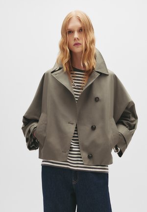 Olivgrüne Doppelt-Mantel-Jacke mit großem Kragen, überschnittenen Schultern, Knopf-Akzenten und Seitentaschen, über einem gestreiften Hemd getragen.