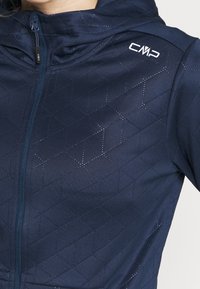 Mörkblå zip-up jacka med ett texturerat geometriskt mönster, med en huva och en liten logotyp på axeln. Slätt material.