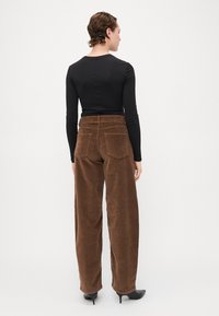 Pantaloni largi din catifea maro, cu un material texturat și buzunare la spate, asortați cu un top negru ajustat cu mânecă lungă și pantofi negri cu vârful ascuțit.