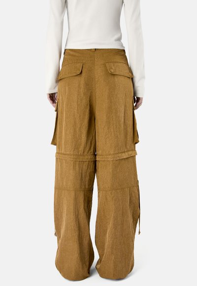 Brązowe szerokie spodnie cargo z wieloma kieszeniami z klapkami i regulowanymi zamkami u dołu, noszone z białą bluzką z długim rękawem.