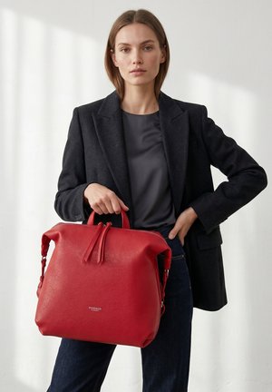 Woman wearing black blazer and dark top holding large red leather handbag with handles, standing against a plain light background.