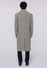 Lungo cappotto grigio di lana con taglio dritto, spacchi posteriori singoli, revers a punta e polsini con bottone, abbinato a pantaloni scuri e scarpe.