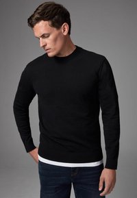 Zwarte gebreide crewneck trui met lange mouwen, geribbelde manchetten en tailleband, gedragen over een wit shirt, gestyled met donkerblauwe jeans.