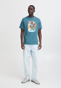 Teal bomulds t-shirt med et farverigt grafisk billede af en abe, der surfer. Sammen med lyseblå jeans og grå sneakers. Simpelt crew neck design.