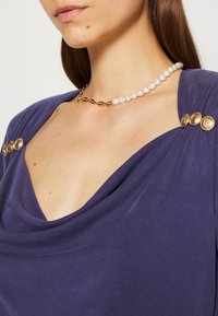 Fialový top s drapérií, který má zlaté knoflíkové akcenty na ramenou a hladkou texturu, doplněný perlovým náhrdelníkem se zlatým řetízkem.