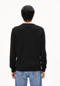 Schwarzer Pullover mit langen Ärmeln, Rundhalsausschnitt, strukturiertem Rückenpatch sowie gerippten Bündchen und Saum. Getragen mit hellblauen Jeans.