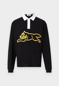 Μαύρο πόλο με μακριά μανίκια και λευκό γιακά, με μεγάλο κίτρινο γραφικό απεικόνιση ενός πλάσματος που πηδά με υπερβολικά χαρακτηριστικά στο μπροστινό μέρος.