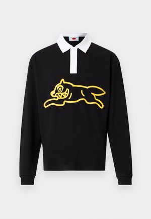 Fekete hosszú ujjú galléros póló fehér gallérral, elöl nagy sárga grafikával, amely egy ábrázolt, túlzó vonásokkal rendelkező ugró lényt ábrázol.