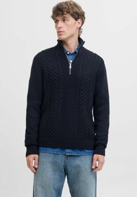 Suéter de punto azul marino con cuello alto y cremallera de un cuarto, con patrones texturizados y puños acanalados. Lleva puesto sobre una camisa de mezclilla.