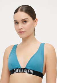 Teal bikiniöverkropp med djup urringning, tillverkad av slät tyg. Har ett svart elastiskt band med "CALVIN KLEIN" logotyp i vitt.